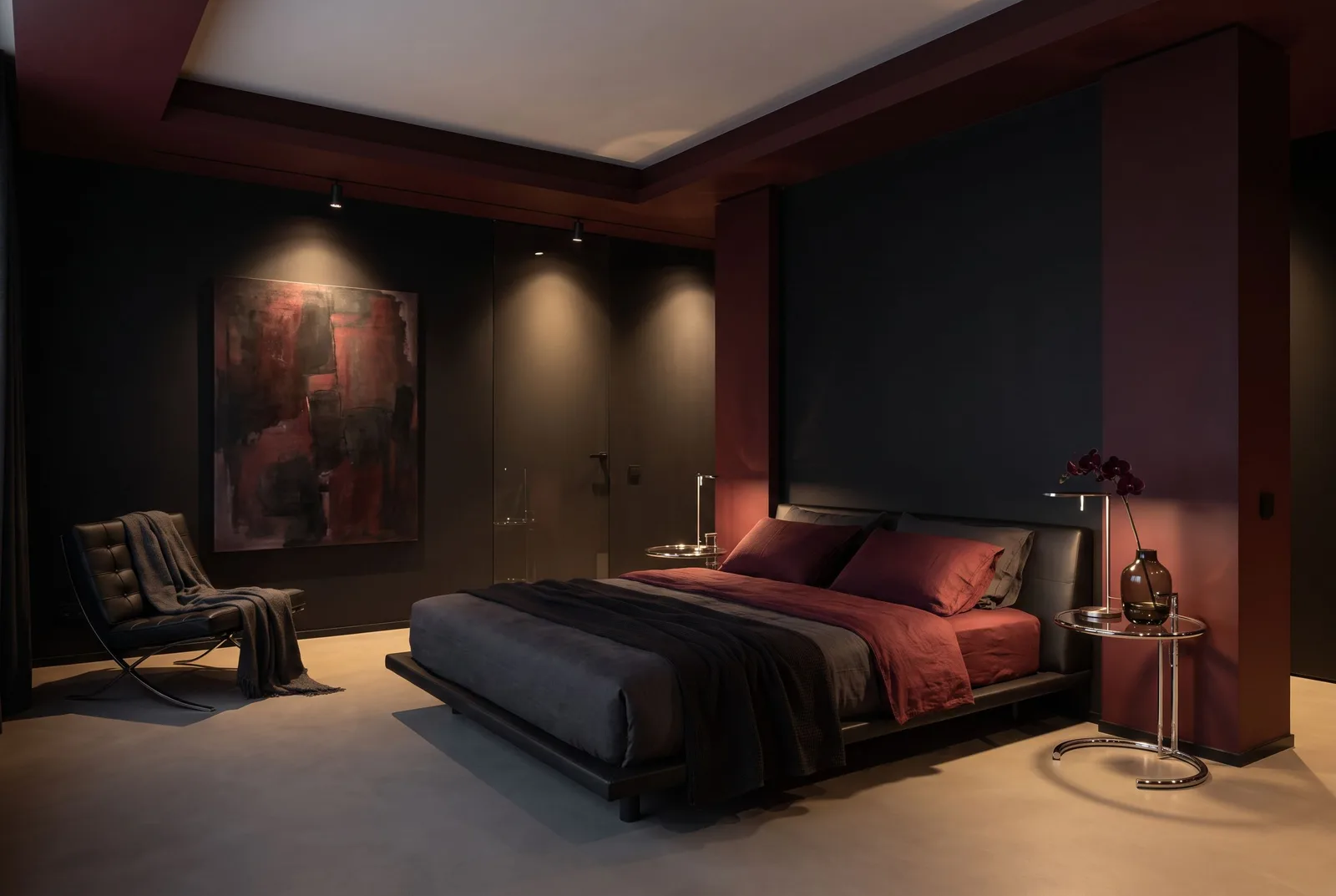 A Modern Noir bedroom with matte black and oxblood walls, oxblood linen on a low leather bed, gallery spotlights on a dark abstract painting