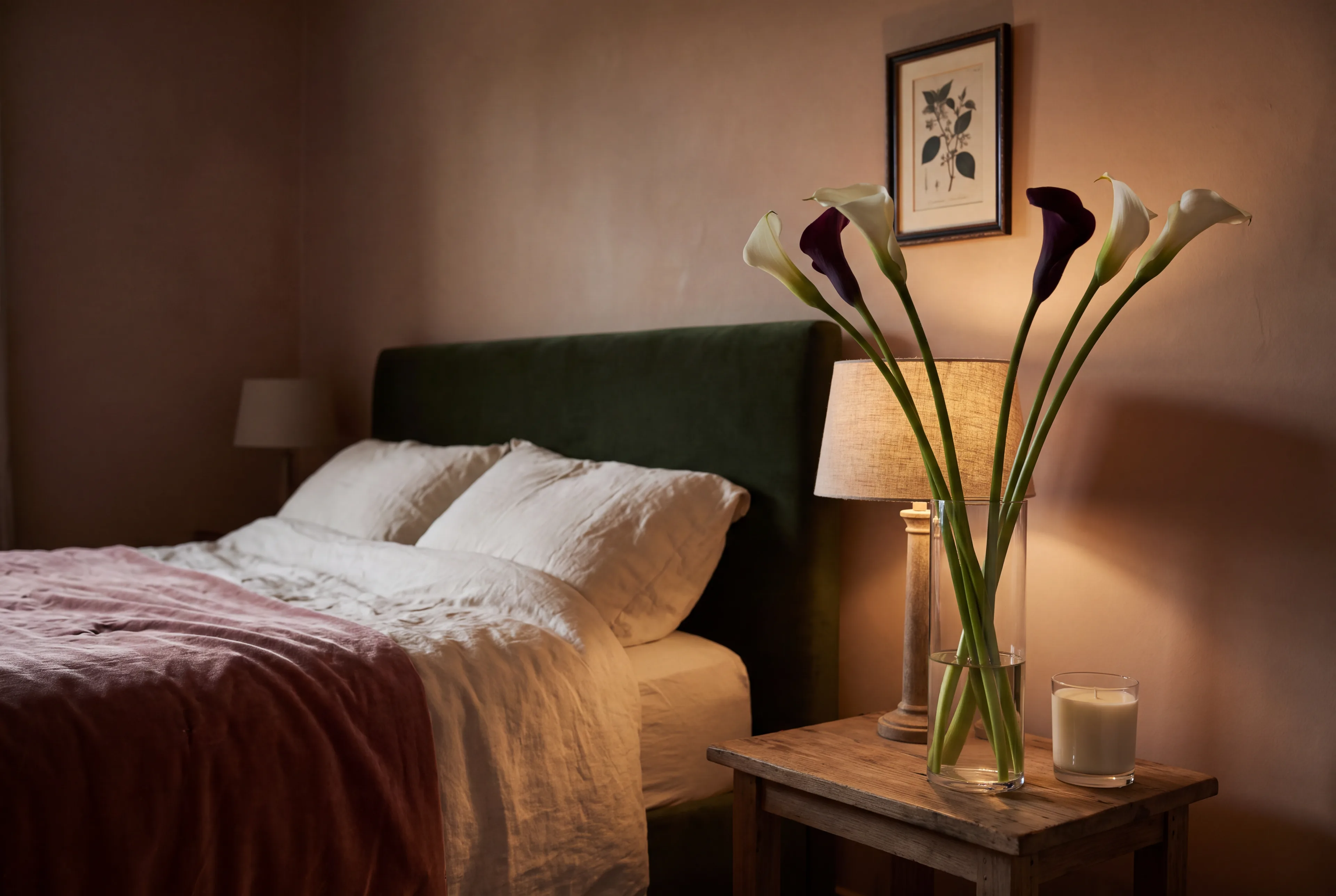 White and black calla lilies in a tall glass vase on a lamplit nightstand beside a linen bed
